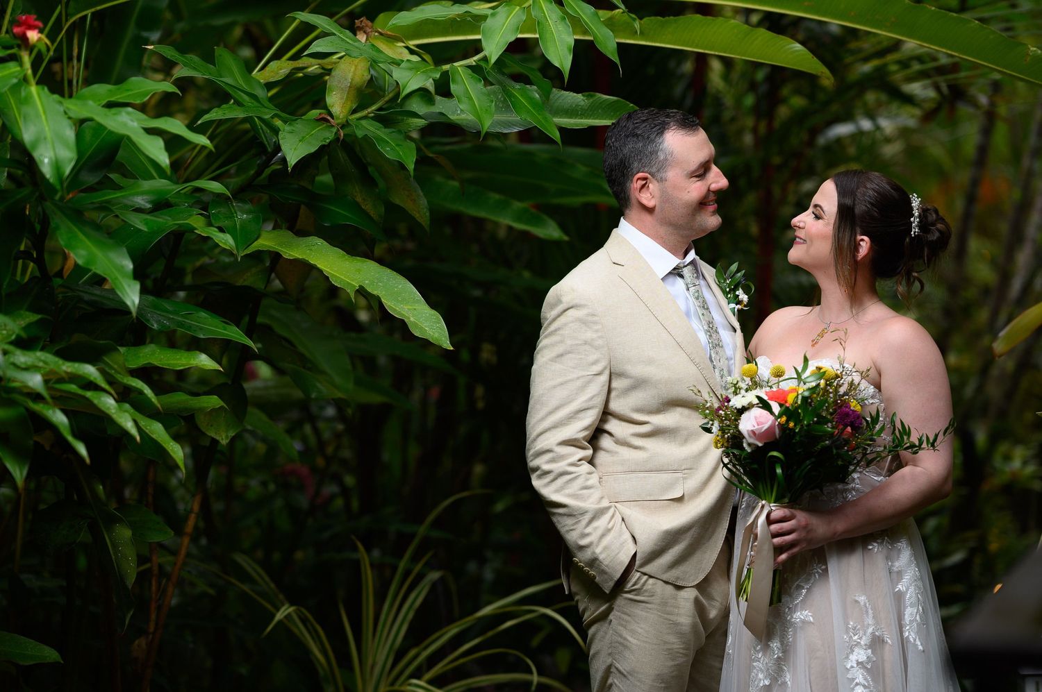 Bride and groom share a romantic moment surrounded by lush tropical greenery, bride holding colorful bouquet.