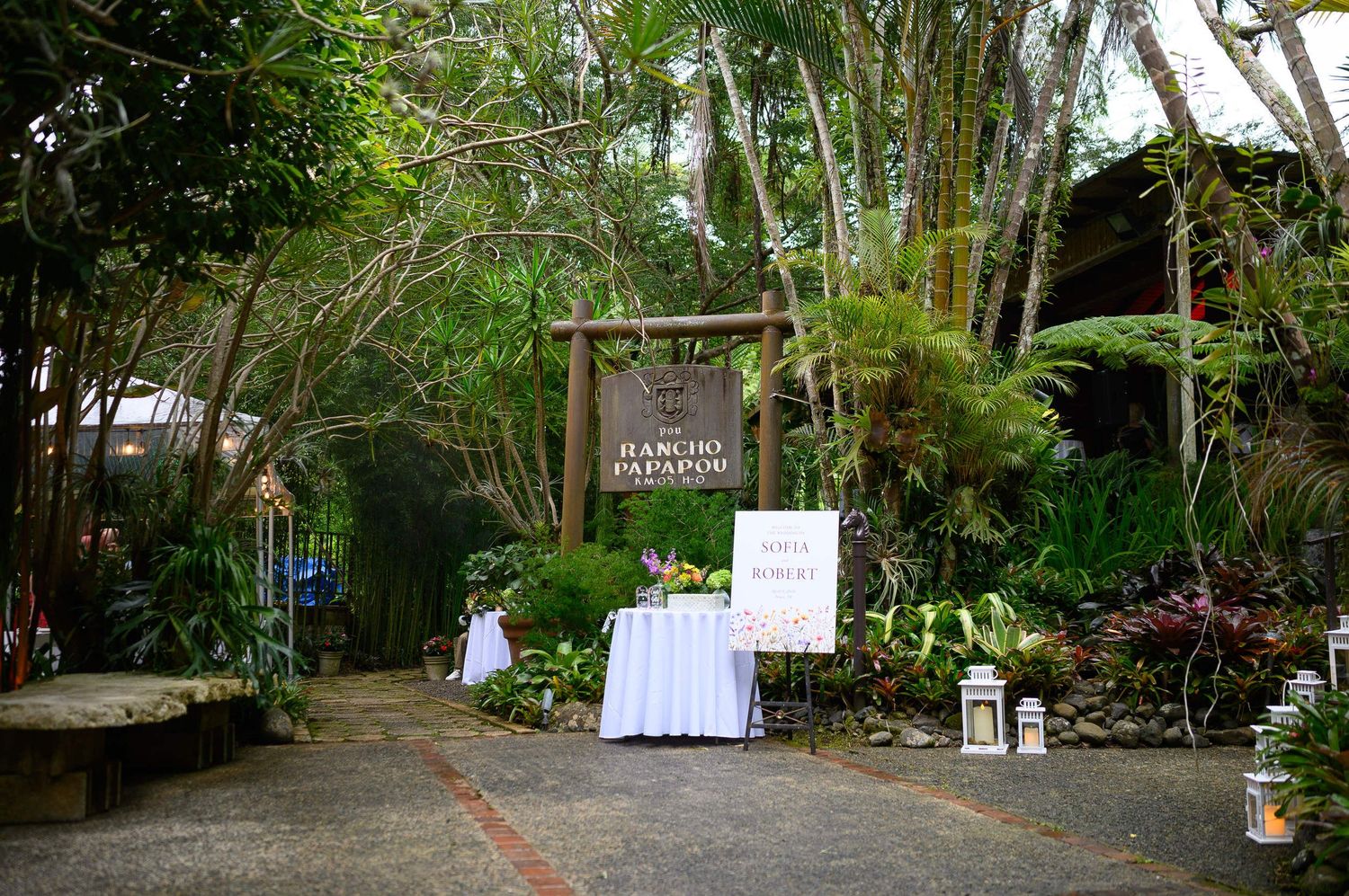 Lush tropical garden entrance with wooden sign, white draped tables, and lanterns at an outdoor venue.