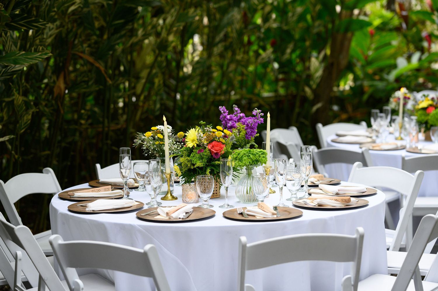 Elegant outdoor wedding reception table with white linens, gold candlesticks, and colorful floral centerpiece.