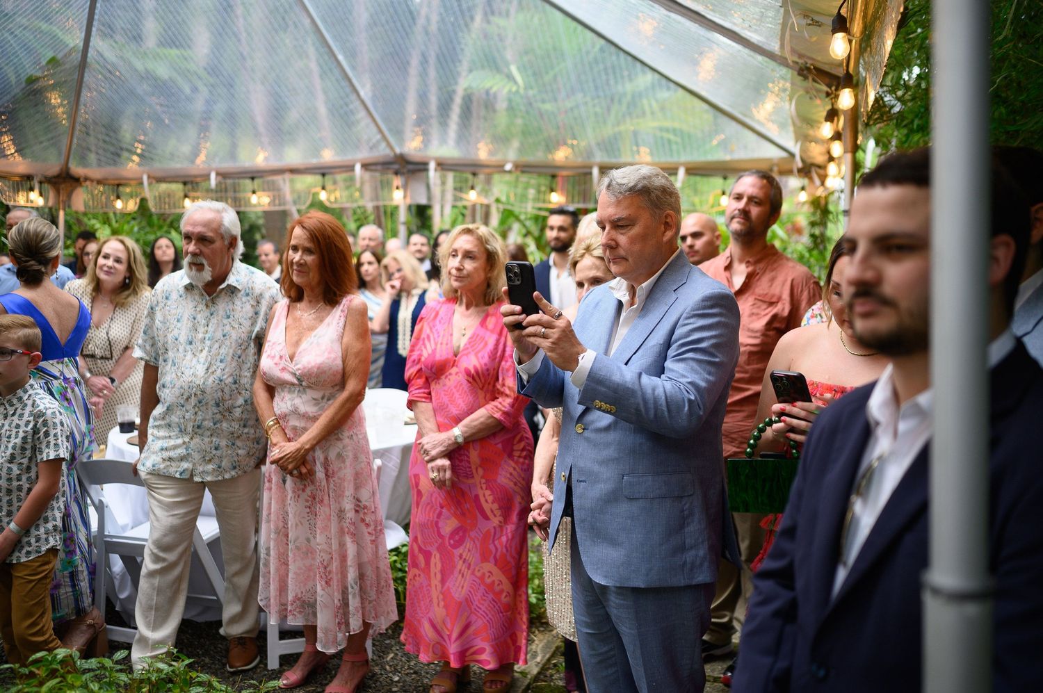 Guests gathered under a transparent tent at an elegant outdoor event, dressed in formal attire amid lush greenery.