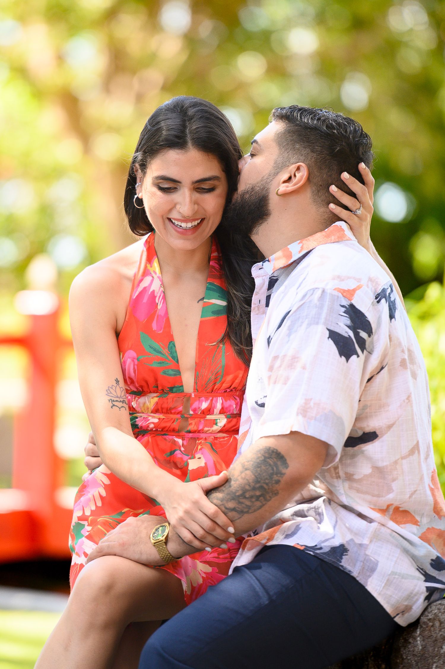 A couple shares a romantic moment outdoors, woman in floral dress laughing as man whispers in her ear.