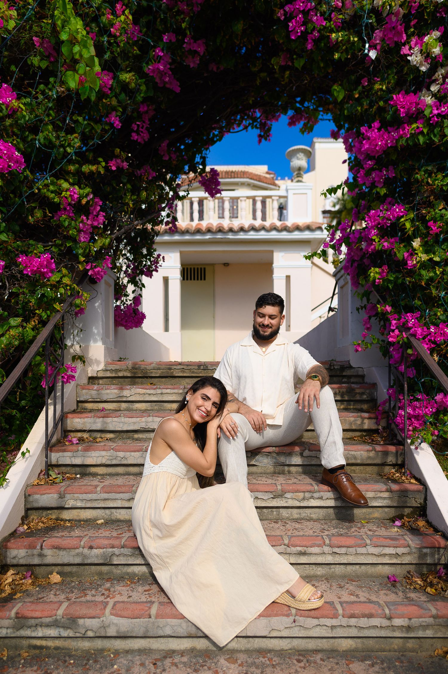 Couple posing on brick steps framed by vibrant pink bougainvillea with a white Mediterranean building behind them.