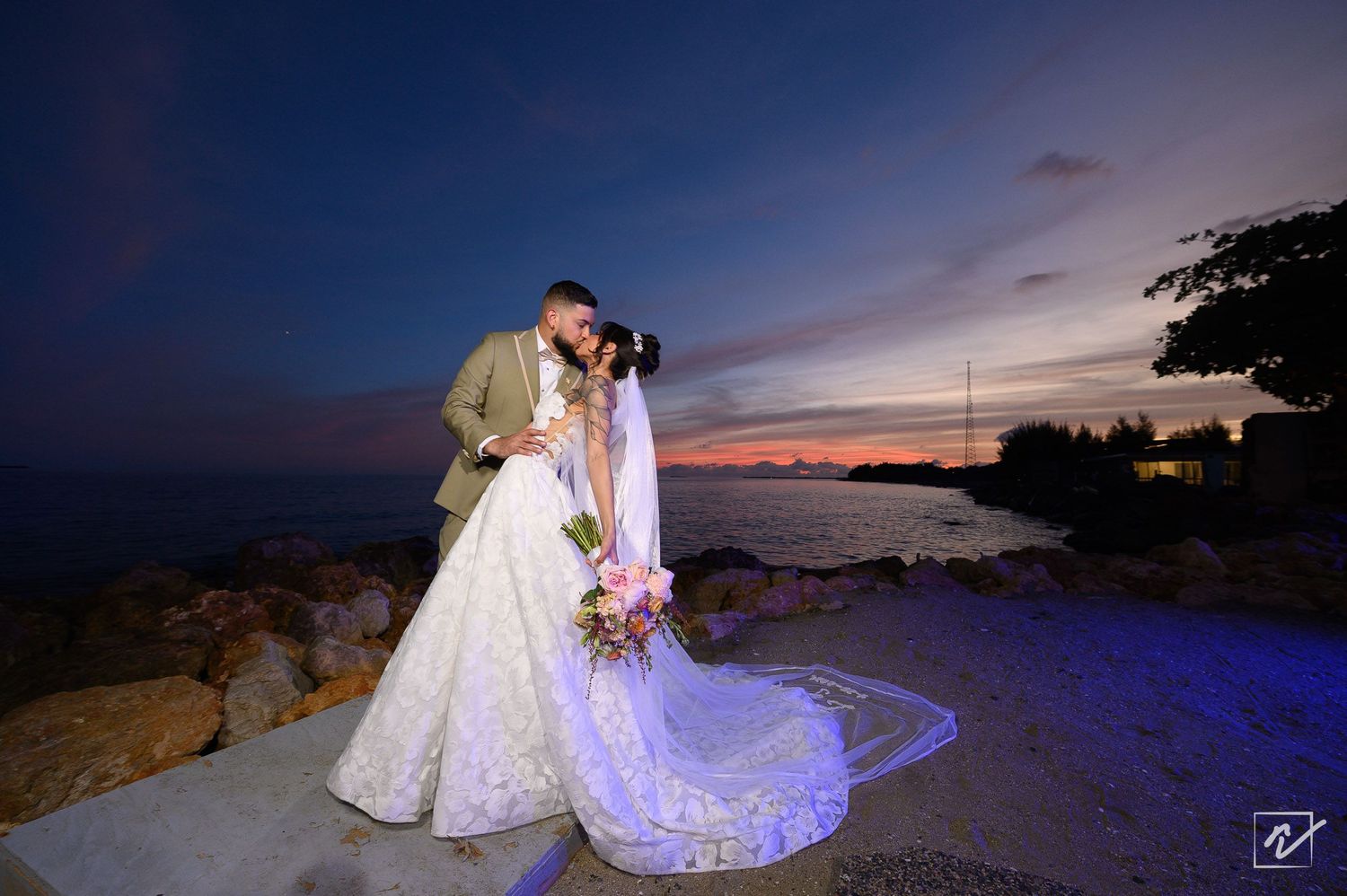 Bride in a flowing white gown holding a bouquet kisses her groom on a rocky seaside shore at sunset under a deep blue twilight sky.