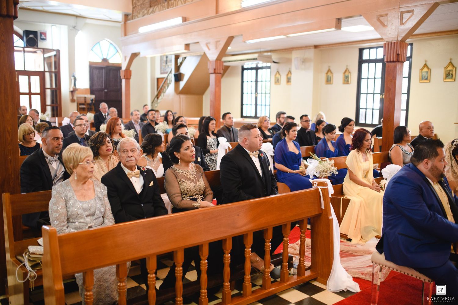 Guests seated in wooden church pews during a wedding ceremony in a bright, decorated Catholic church.