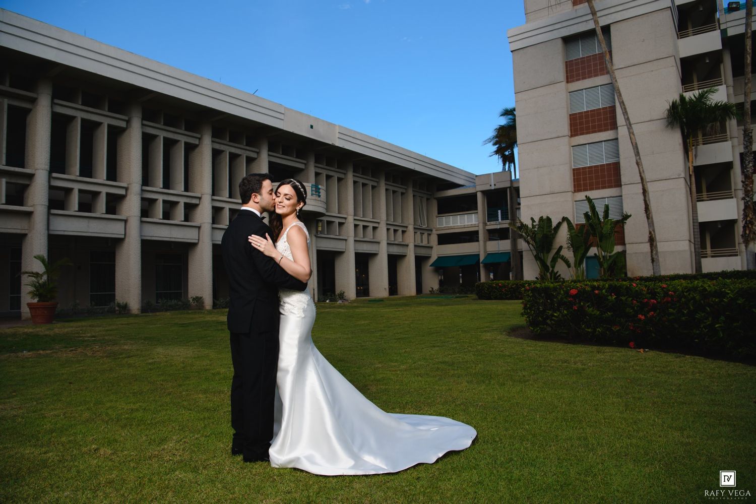 Bride and groom embracing on green lawn in front of modern building on sunny wedding day.