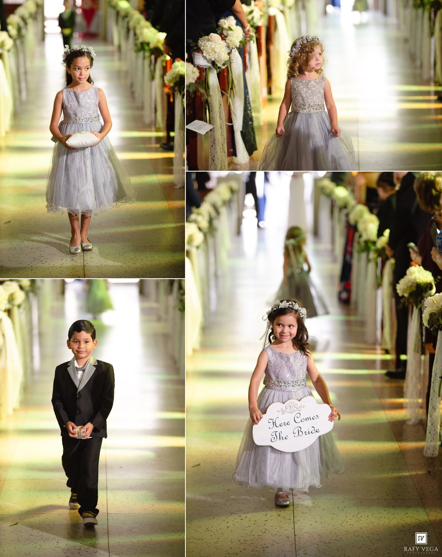 Flower girls and ring bearer walk down church aisle in gray formal attire, one girl holding a Here Comes the Bride sign.