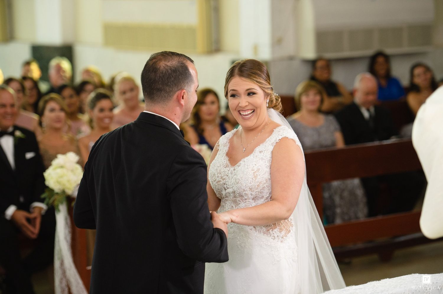 Bride smiling at groom during wedding ceremony in church with guests seated in pews behind them.
