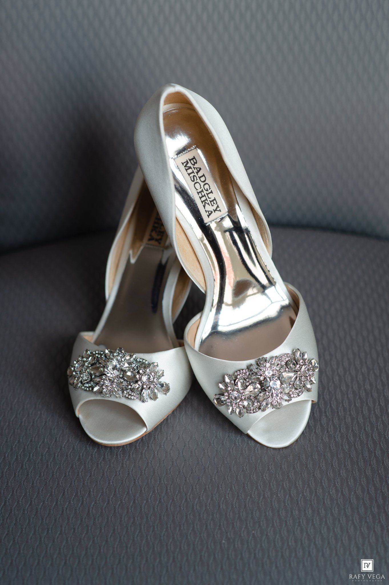 Badgley Mischka white satin peep-toe bridal heels with crystal embellishments displayed on a gray chair.