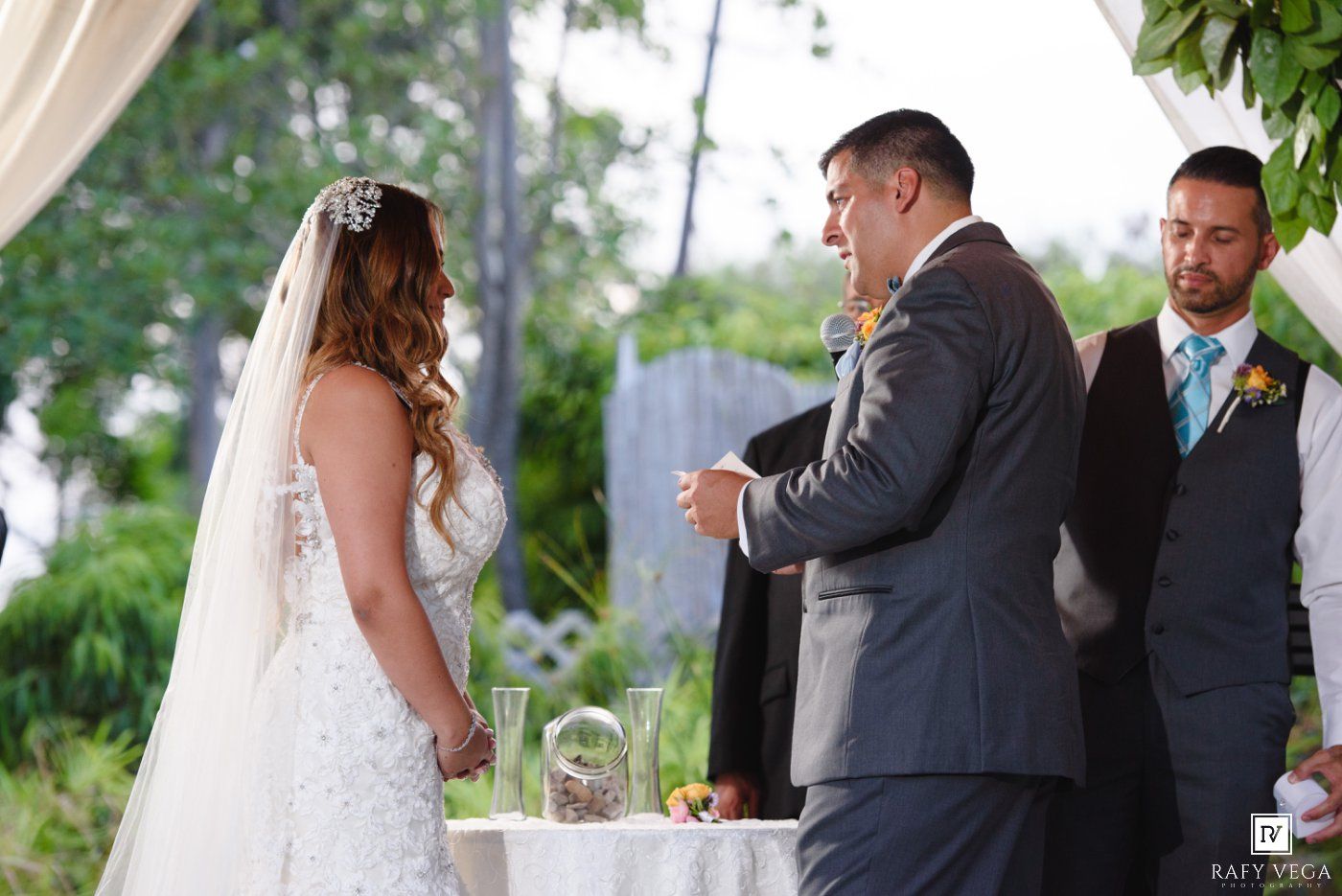 Bride and groom exchange vows at outdoor wedding ceremony with officiant and groomsman in background.