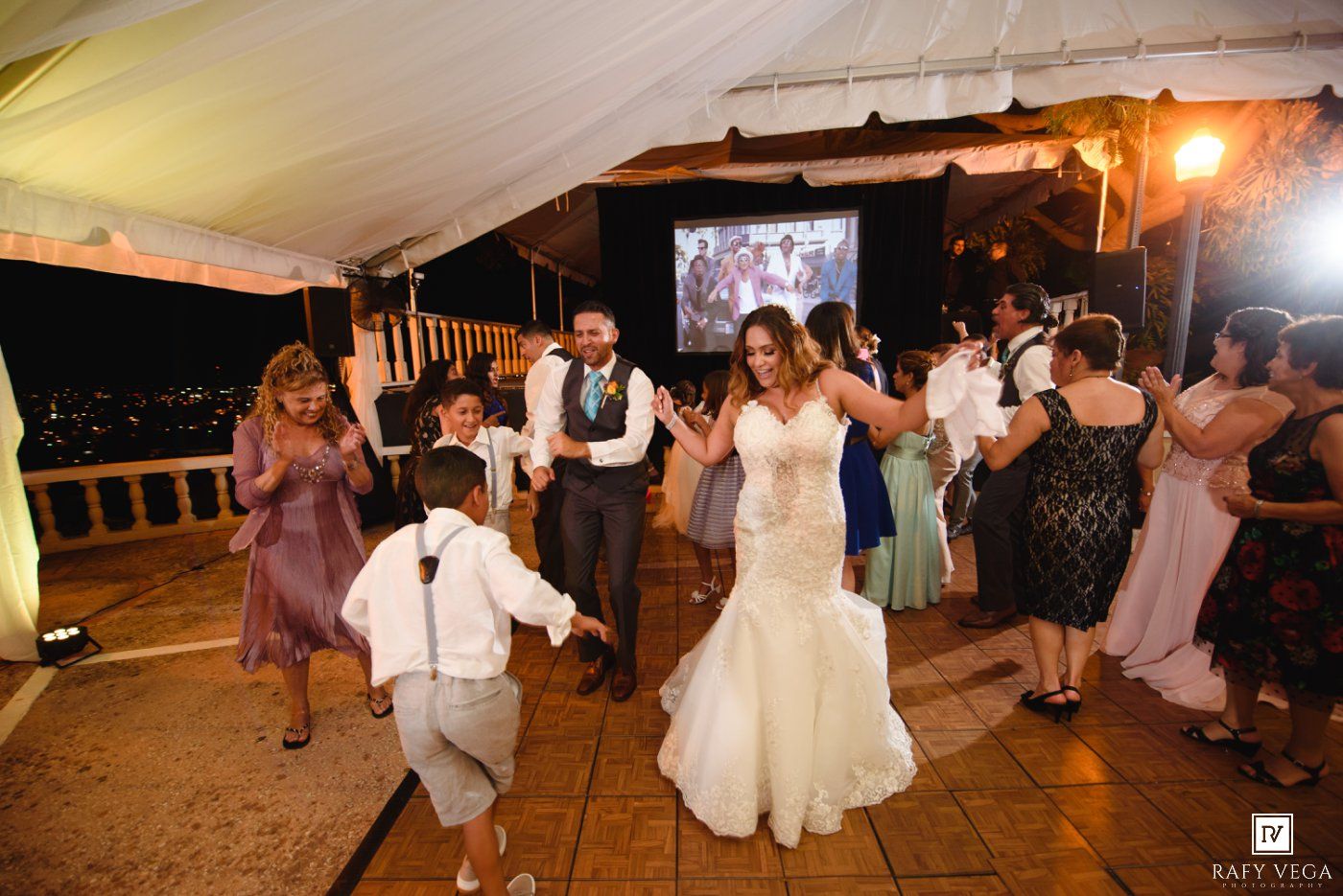Bride and groom dancing joyfully with guests under a tent at an outdoor wedding reception at night.