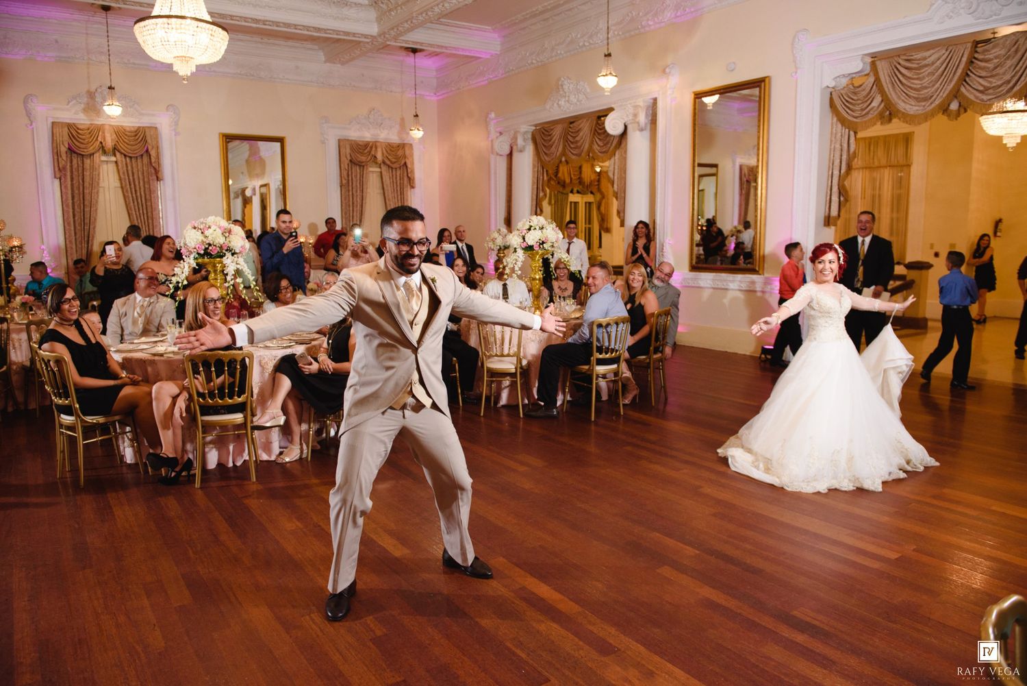 Bride and groom share first dance in elegant ballroom with guests seated at decorated tables during wedding reception.