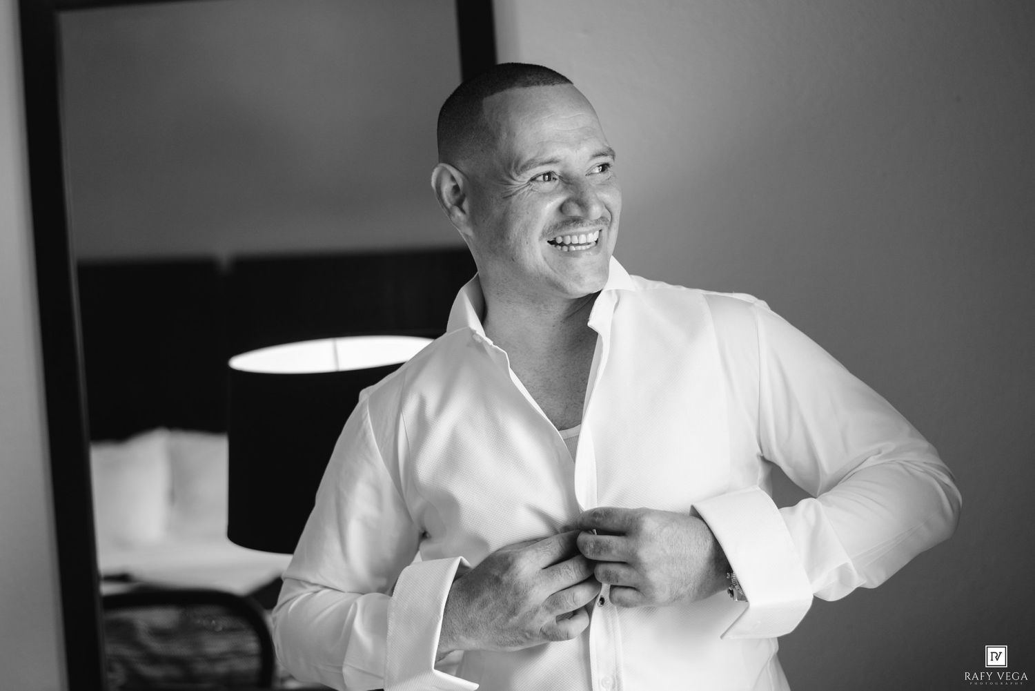 Man in white dress shirt smiling while buttoning up in a hotel room, black and white wedding preparation photo.