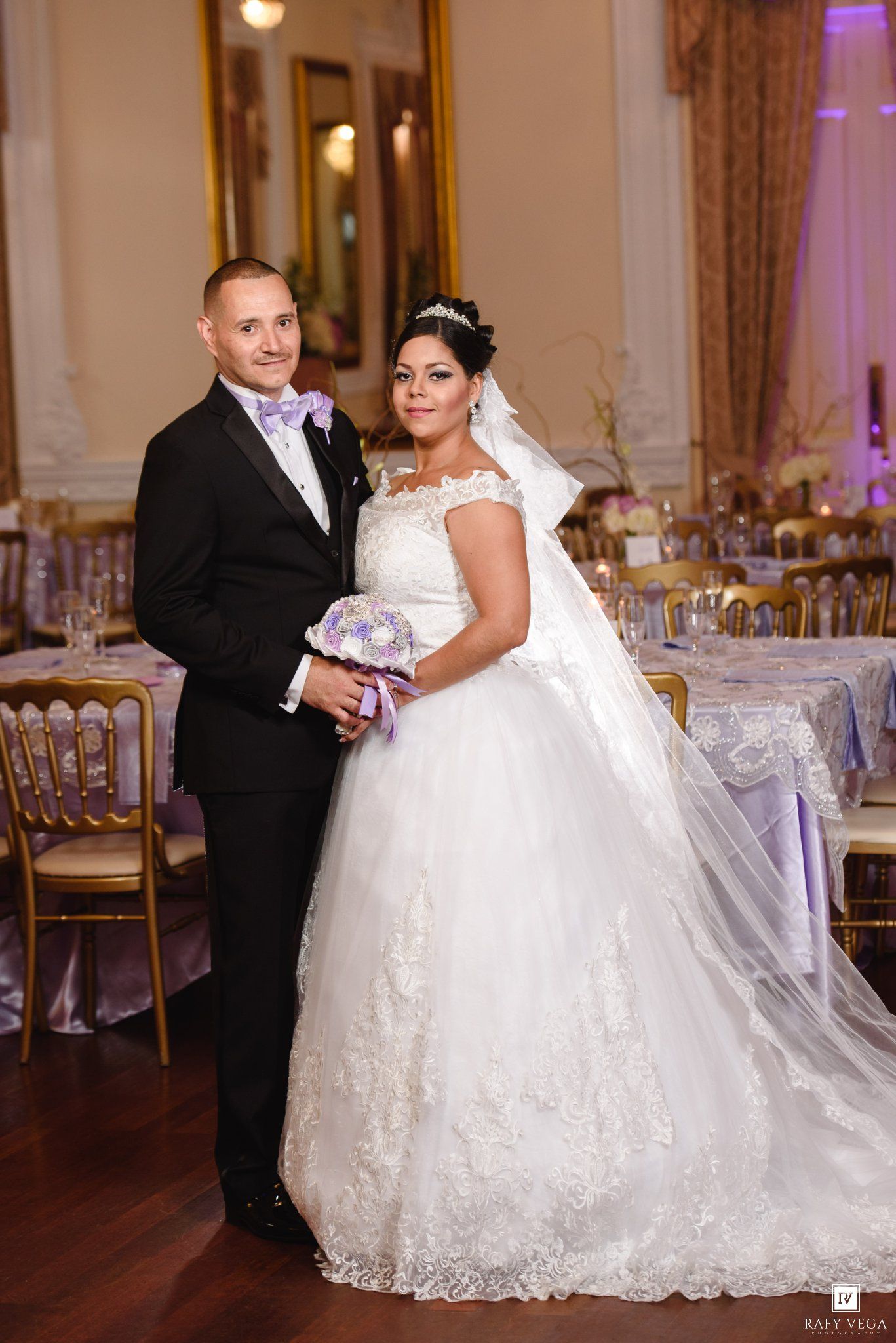 Bride in white lace ball gown and groom in black tuxedo pose together at elegant wedding reception venue.