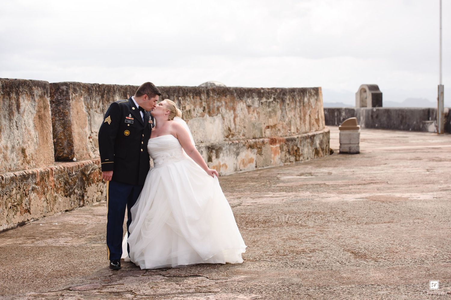 Military groom in dress uniform kissing bride in white wedding gown at historic stone fort outdoor ceremony.