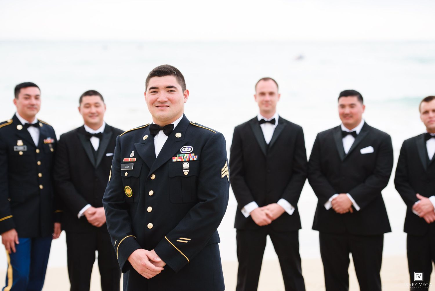 Military groom in Army dress uniform stands with groomsmen in tuxedos on beach wedding day.