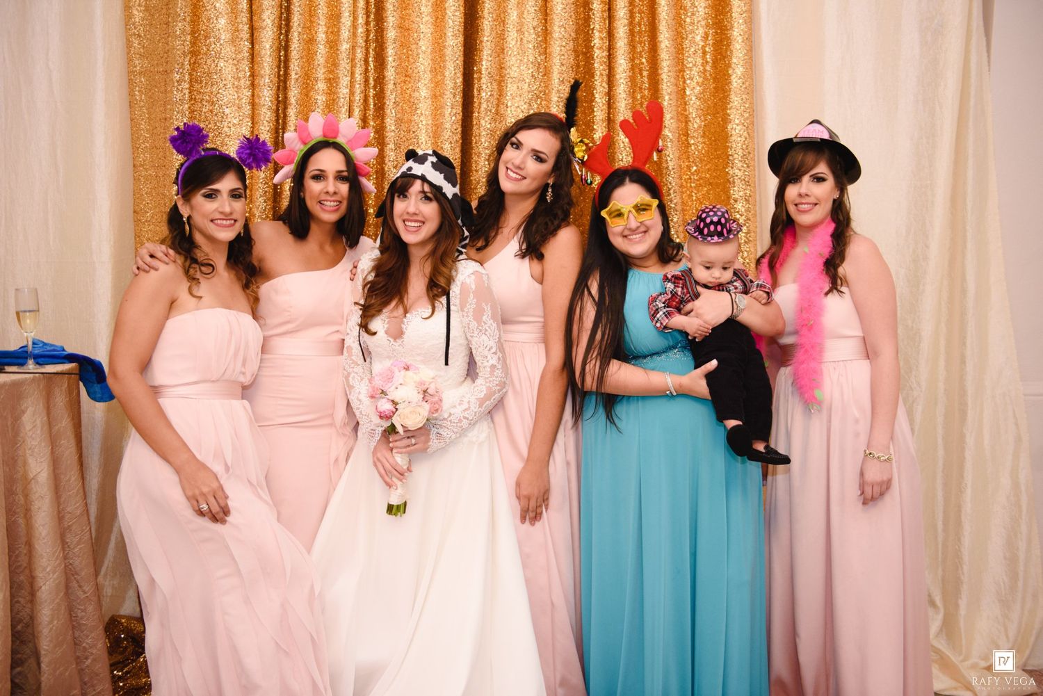 Bride in white gown poses with bridesmaids in pink and blue dresses, wearing fun photo booth props at wedding reception.