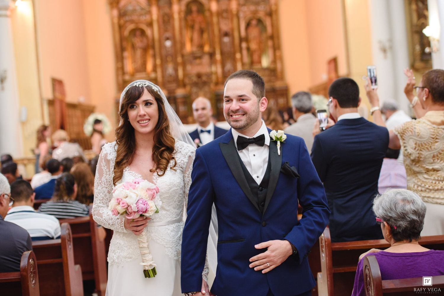 Bride and groom walk down church aisle smiling, bride holding pink bouquet, groom in navy suit with bow tie.