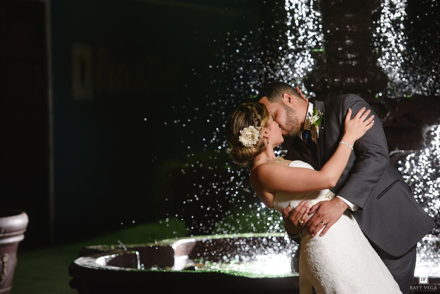 Bride and groom sharing a romantic kiss in front of a sparkling fountain at their elegant outdoor wedding reception.