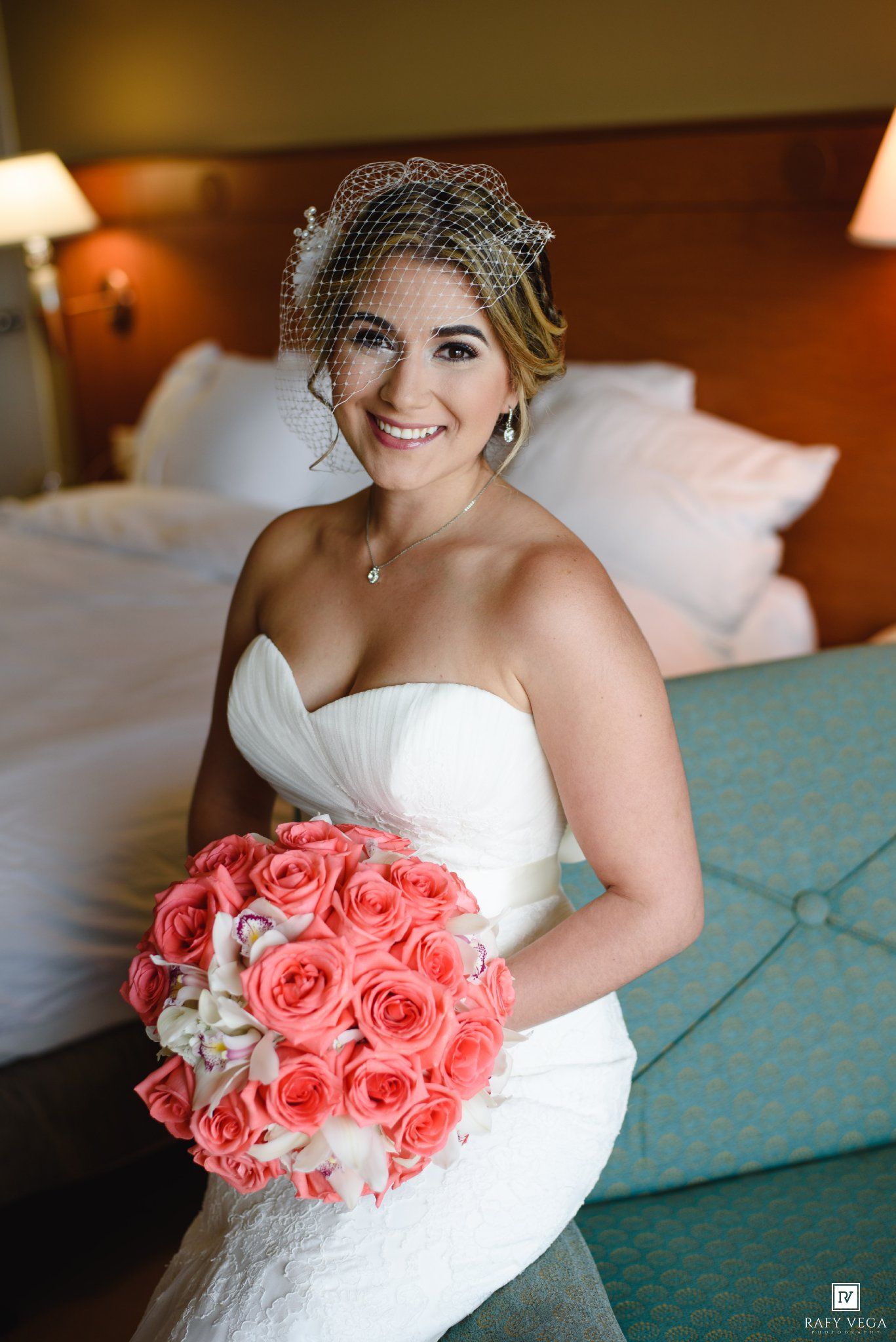 Smiling bride in white strapless gown holding coral rose bouquet, wearing birdcage veil, seated in hotel room.