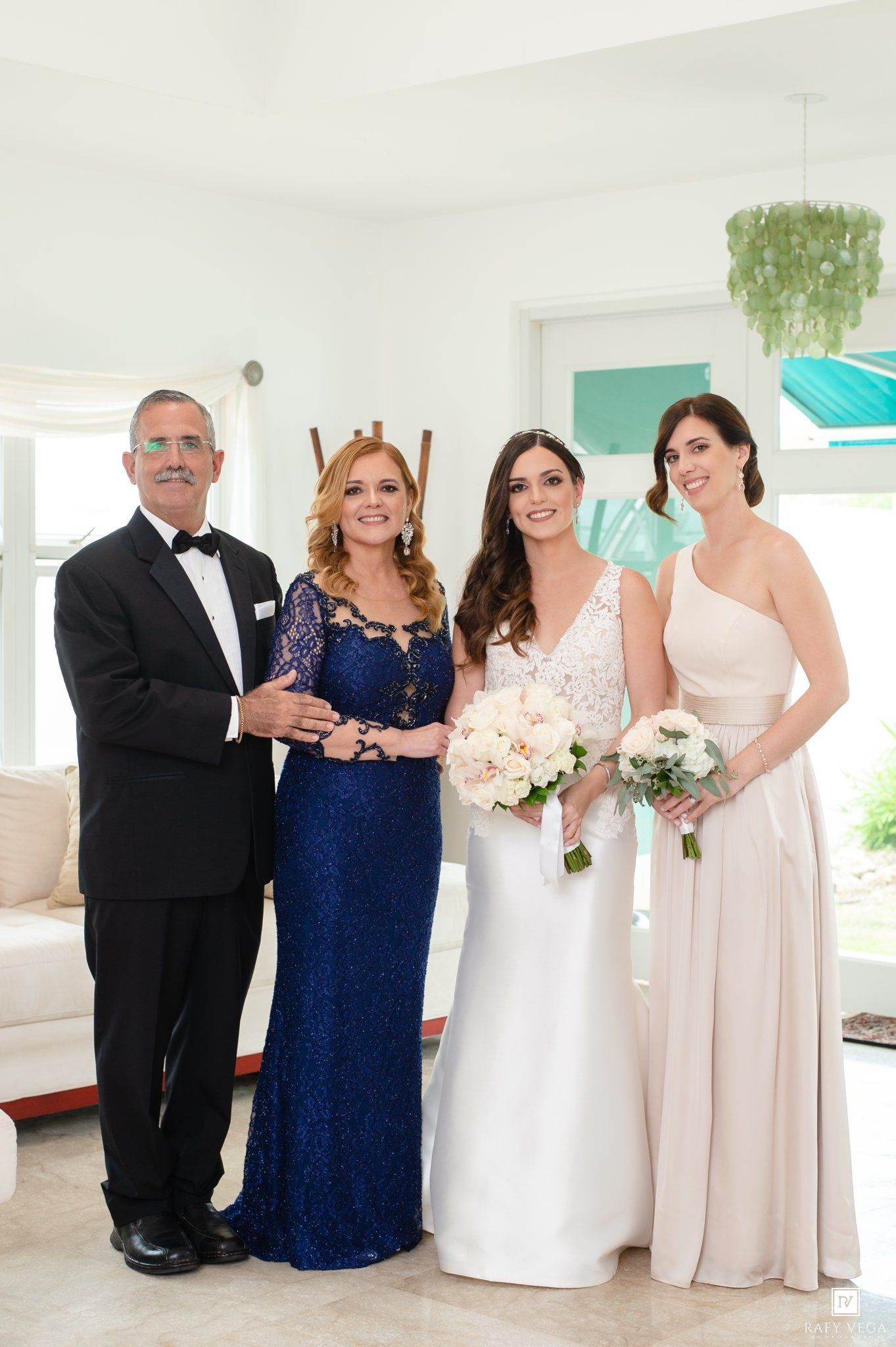 Bride in white gown with bouquet poses alongside bridesmaid and elegantly dressed family members at a bright wedding venue.