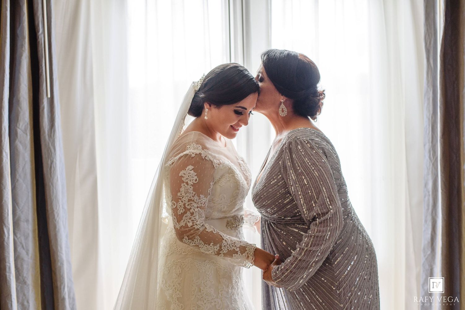 Bride in lace wedding gown shares tender moment with mother in sequined dress by window on wedding day.