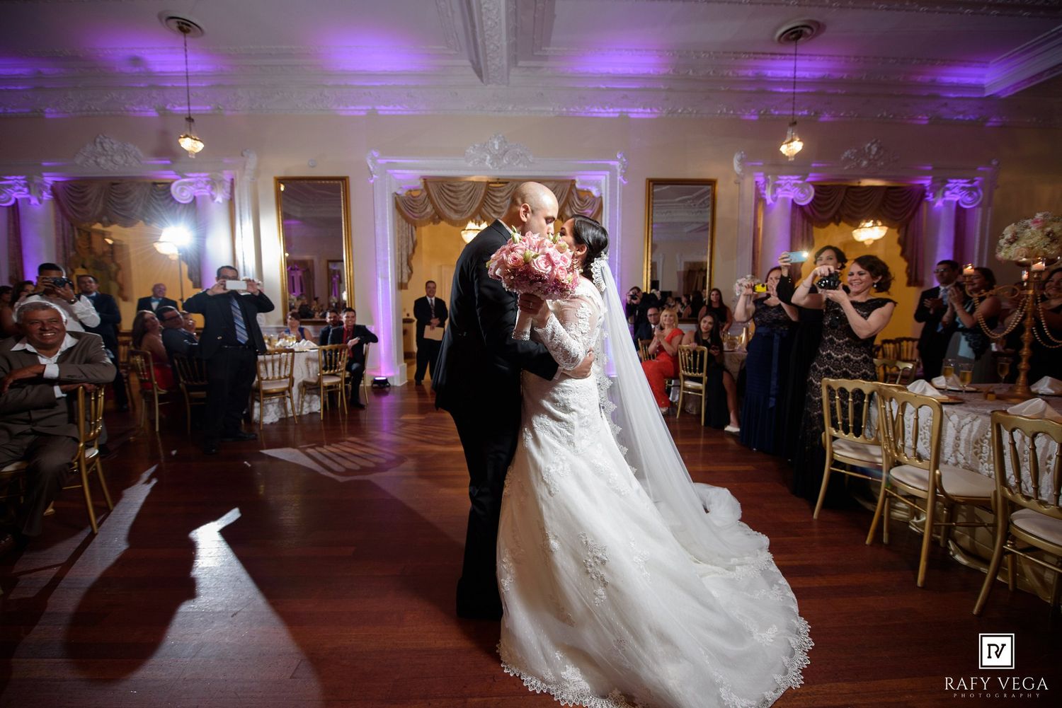Bride and groom share first dance in elegant ballroom with purple uplighting and guests watching at wedding reception.