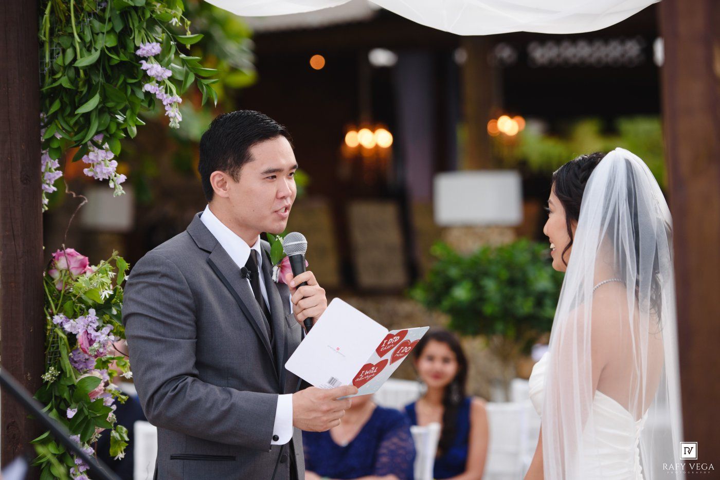 Groom reads wedding vows into microphone at outdoor ceremony, bride in white veil listens, floral arch backdrop.