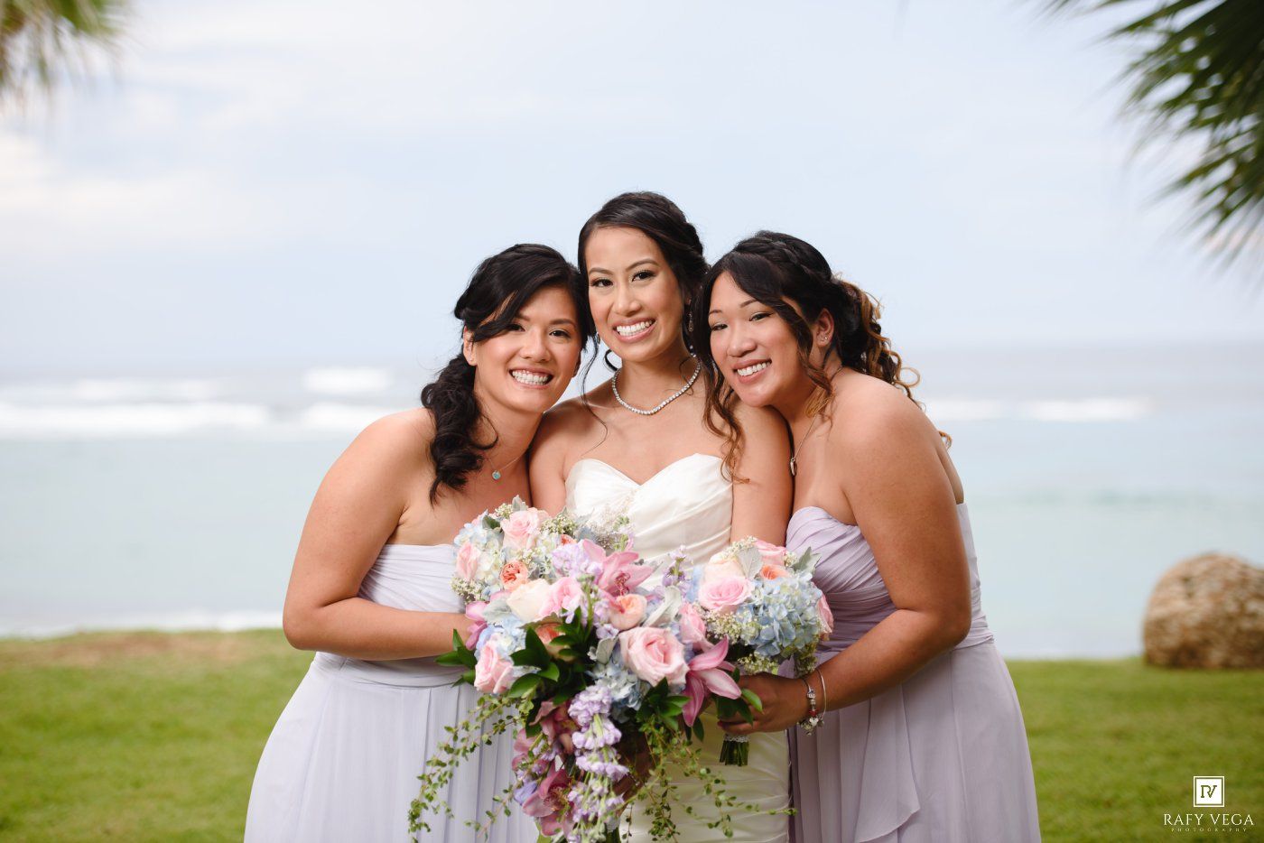 A smiling bride holds a pink bouquet flanked by two bridesmaids in lavender dresses by the ocean.