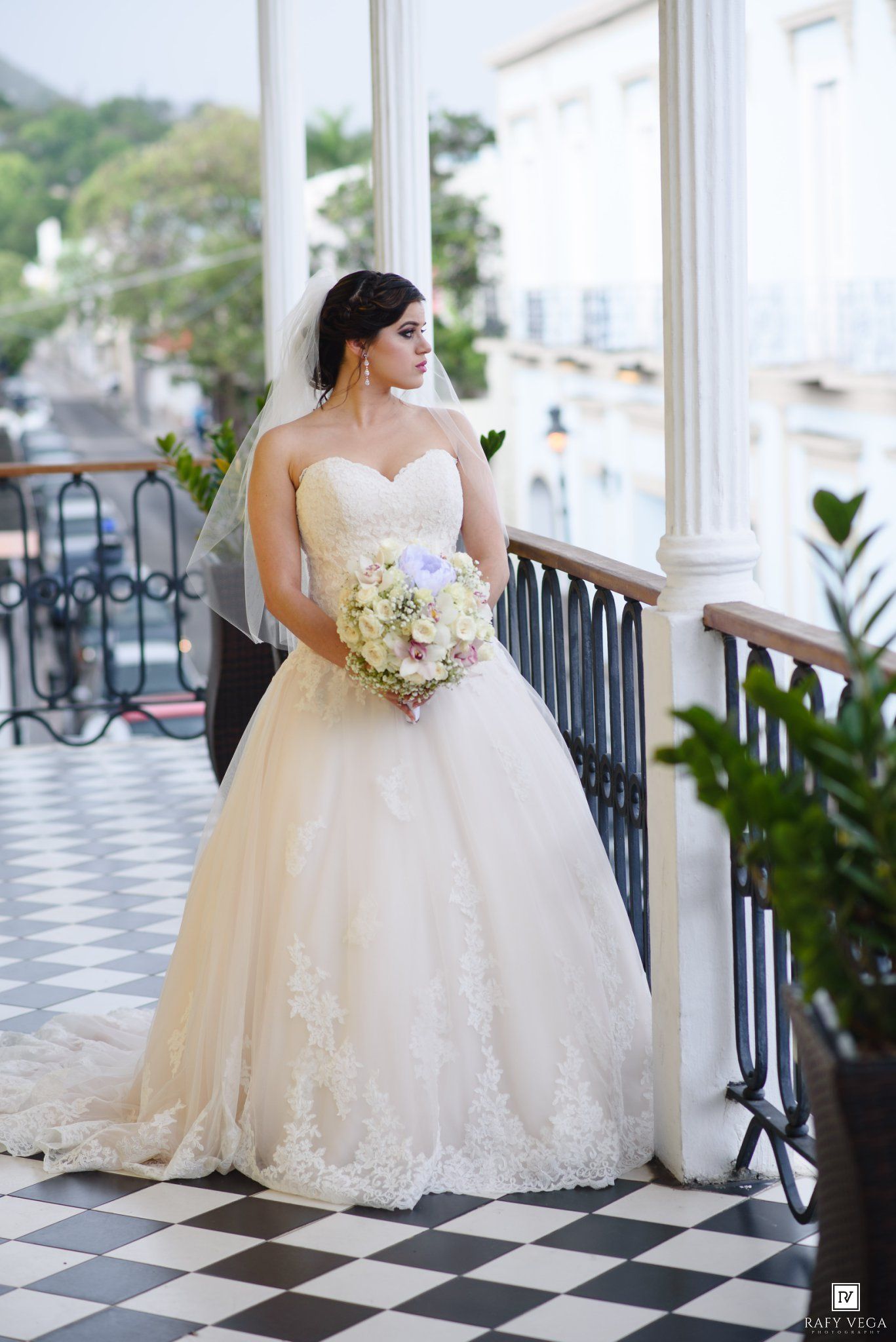 Bride in strapless lace ball gown wedding dress holding floral bouquet on elegant balcony with checkered floor.