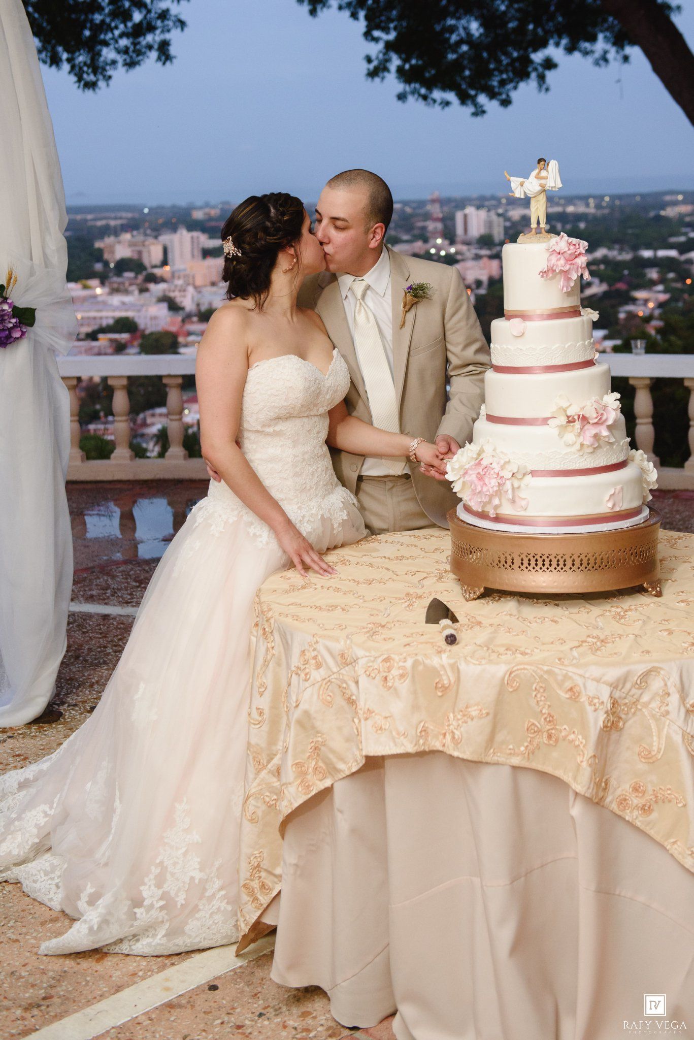 Bride and groom kissing while cutting a four-tier white wedding cake with pink flowers on an outdoor terrace at dusk.