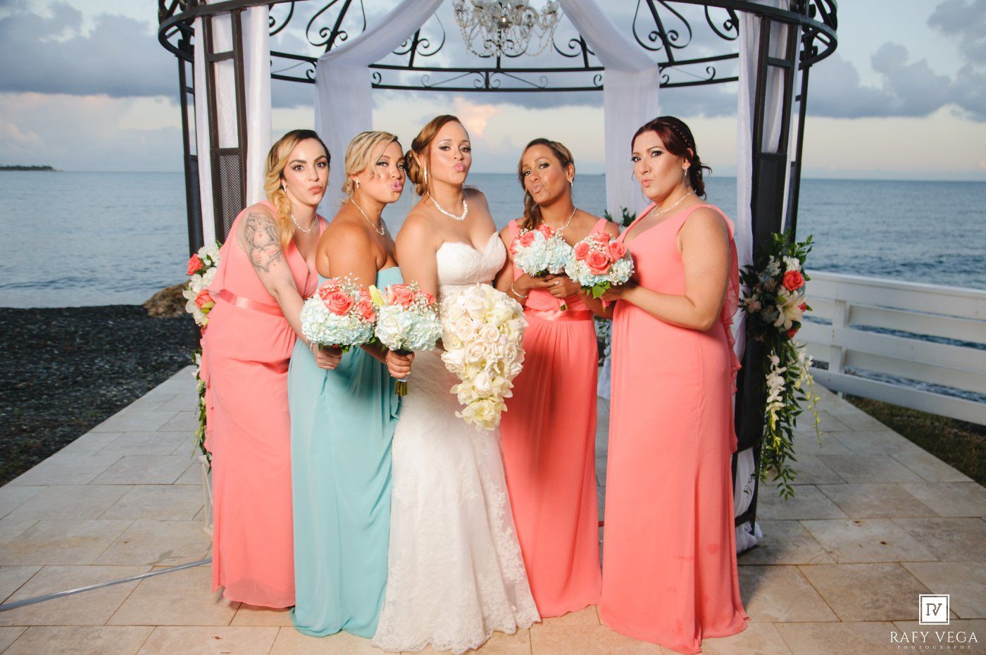 Bride with bridesmaids in coral and mint gowns holding bouquets at seaside gazebo wedding ceremony.