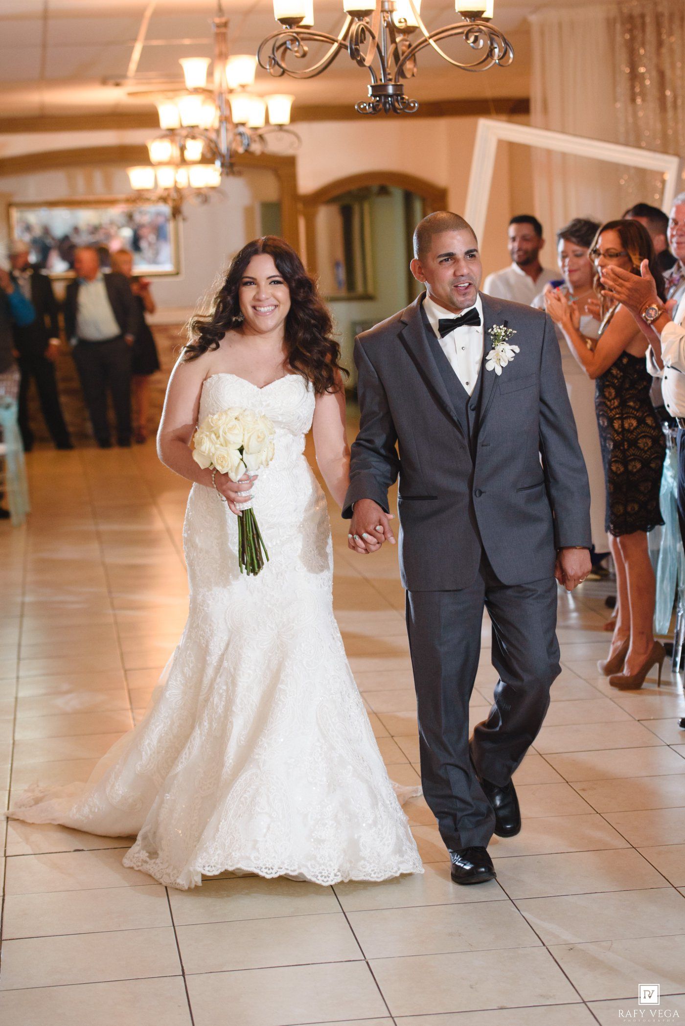 Bride in white lace mermaid gown and groom in gray suit hold hands walking into wedding reception hall.