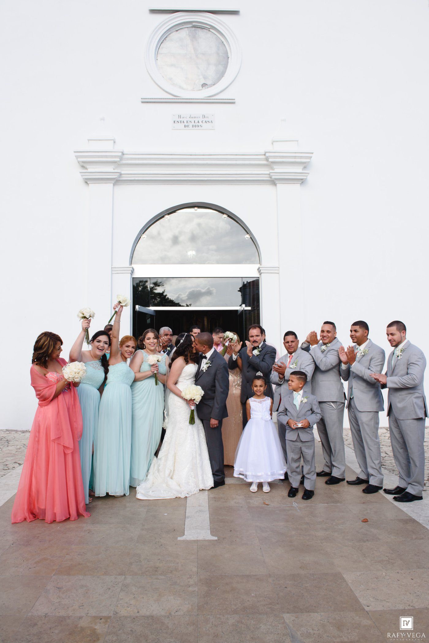 Wedding party poses outside white church, bride in gown, bridesmaids in mint, groomsmen in gray suits, flower girl present.