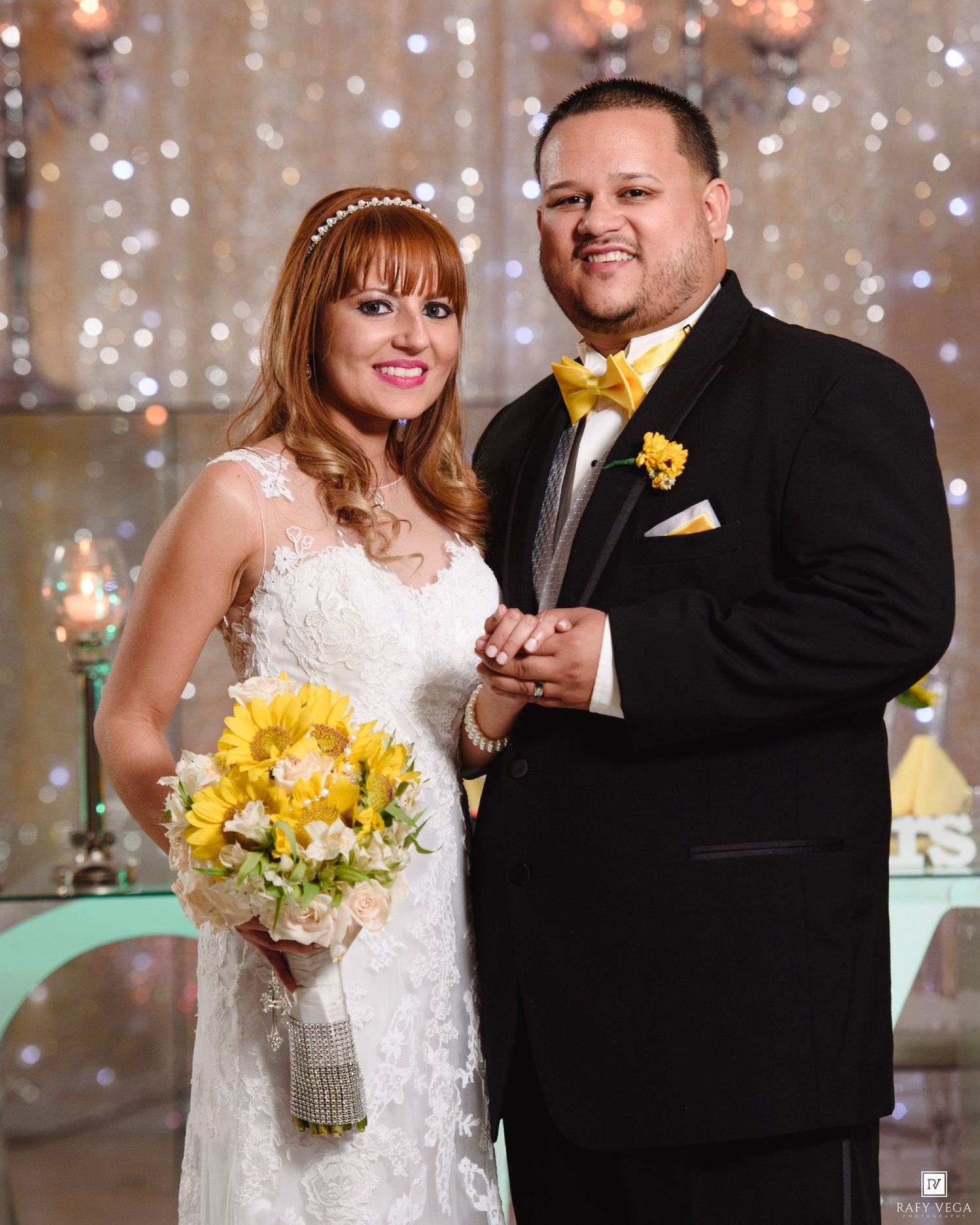 Bride and groom posing together at wedding ceremony, bride holding yellow sunflower bouquet, groom wearing black tuxedo with yellow bow tie.