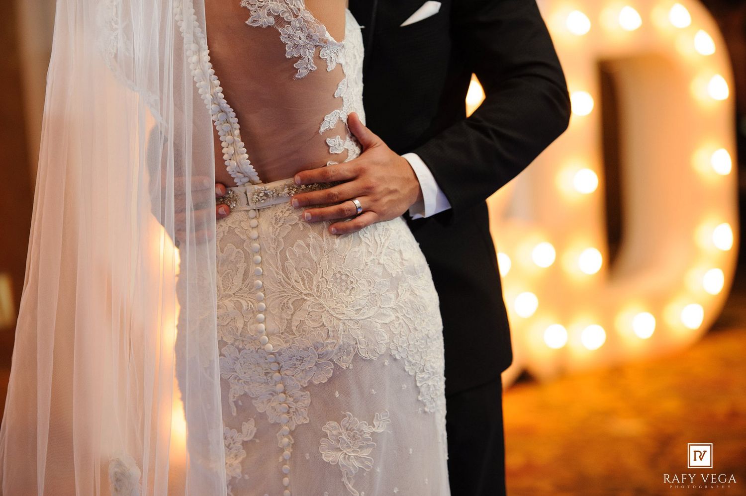 Close-up of bride in lace backless wedding gown with veil, groom in black tuxedo embracing her.