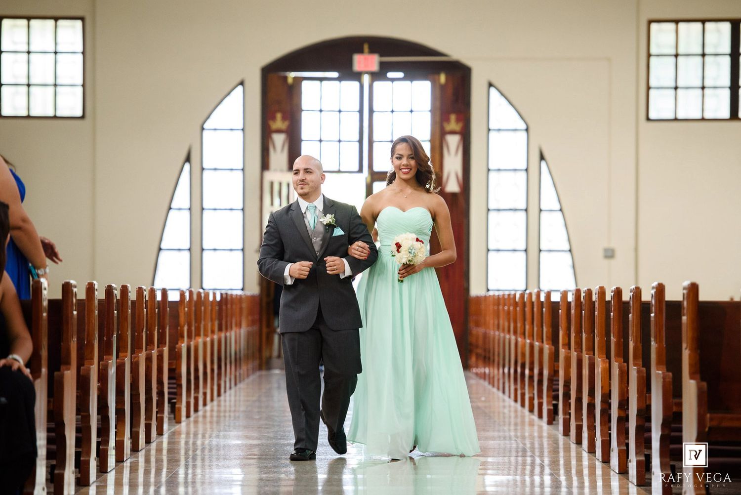 A bridesmaid in mint green gown walks down church aisle with groomsman during elegant wedding ceremony.