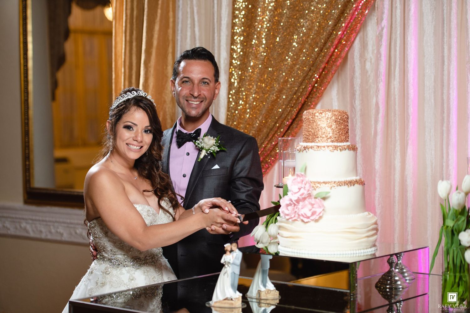 Bride and groom smiling while cutting their elegant white wedding cake at a glamorous gold-themed reception venue.