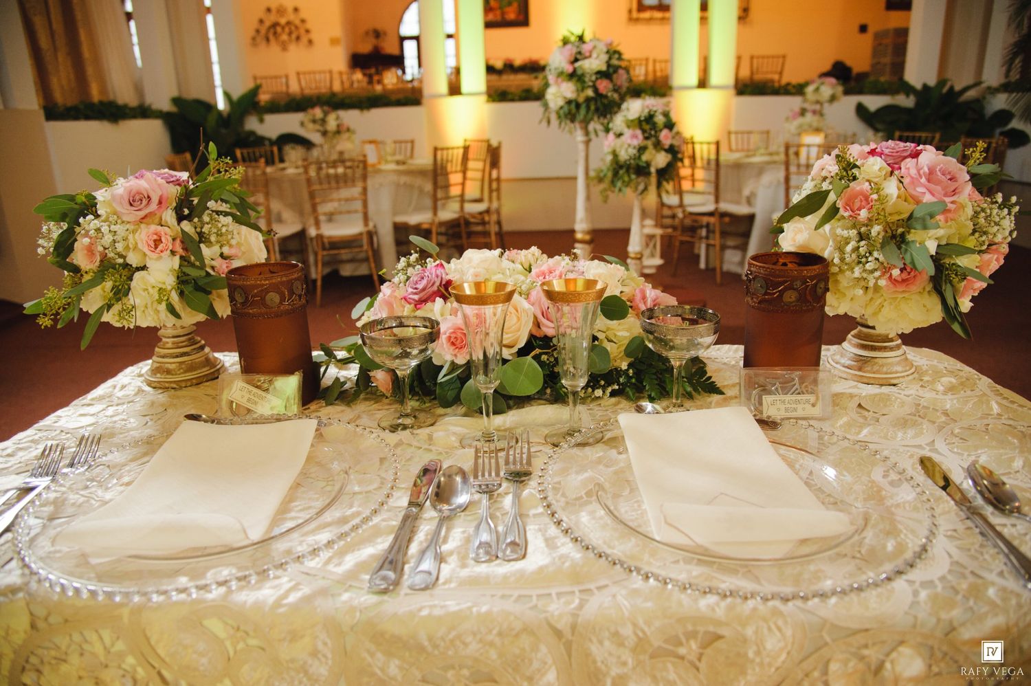Elegant wedding reception table with pink floral centerpieces, gold lace tablecloth, and formal place settings.