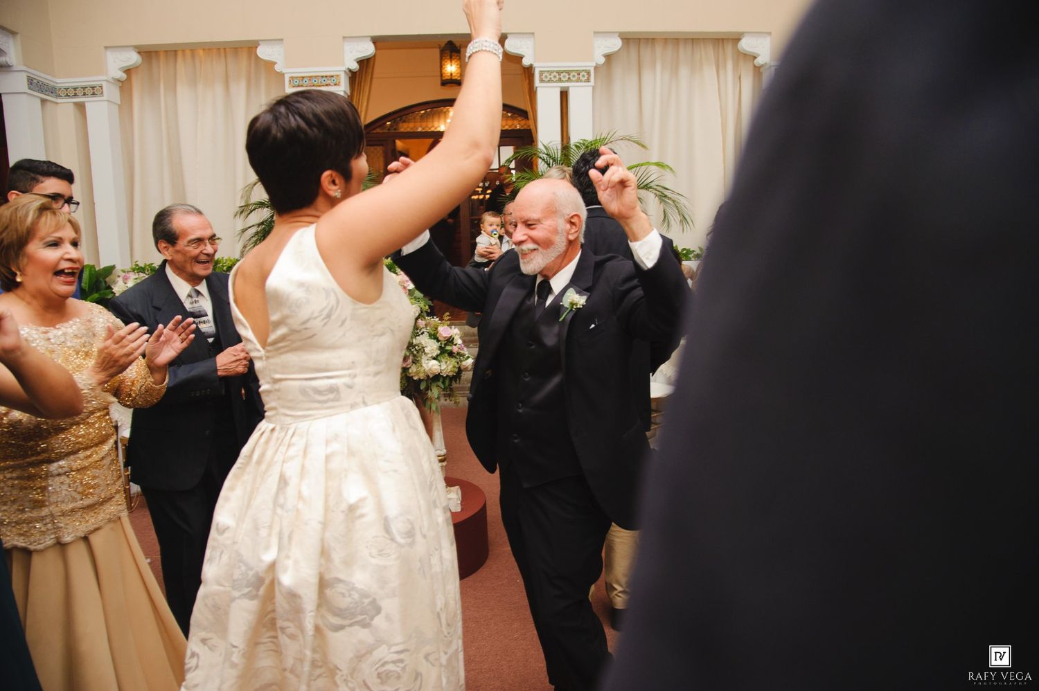 Bride and elderly man dancing joyfully at wedding reception while guests clap and celebrate in elegant venue.