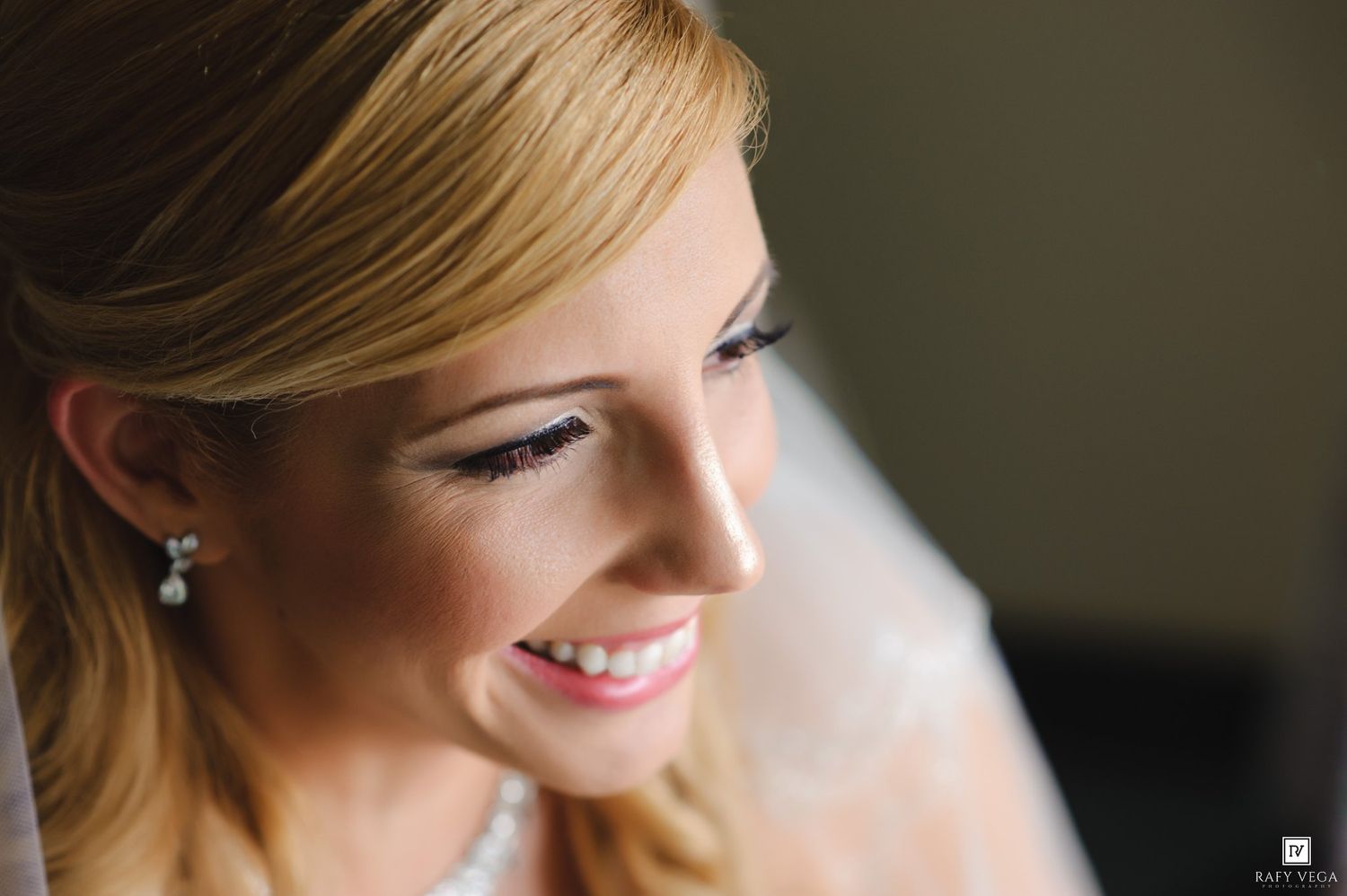 Smiling bride with elegant pearl earrings and veil, showcasing natural bridal makeup with soft pink lips and defined eyes.