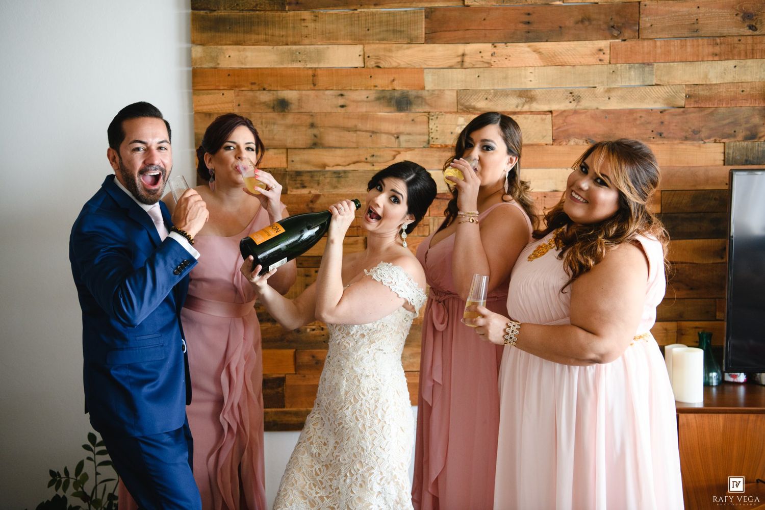 Bride and bridesmaids in blush pink dresses celebrate with champagne bottle before wedding ceremony.