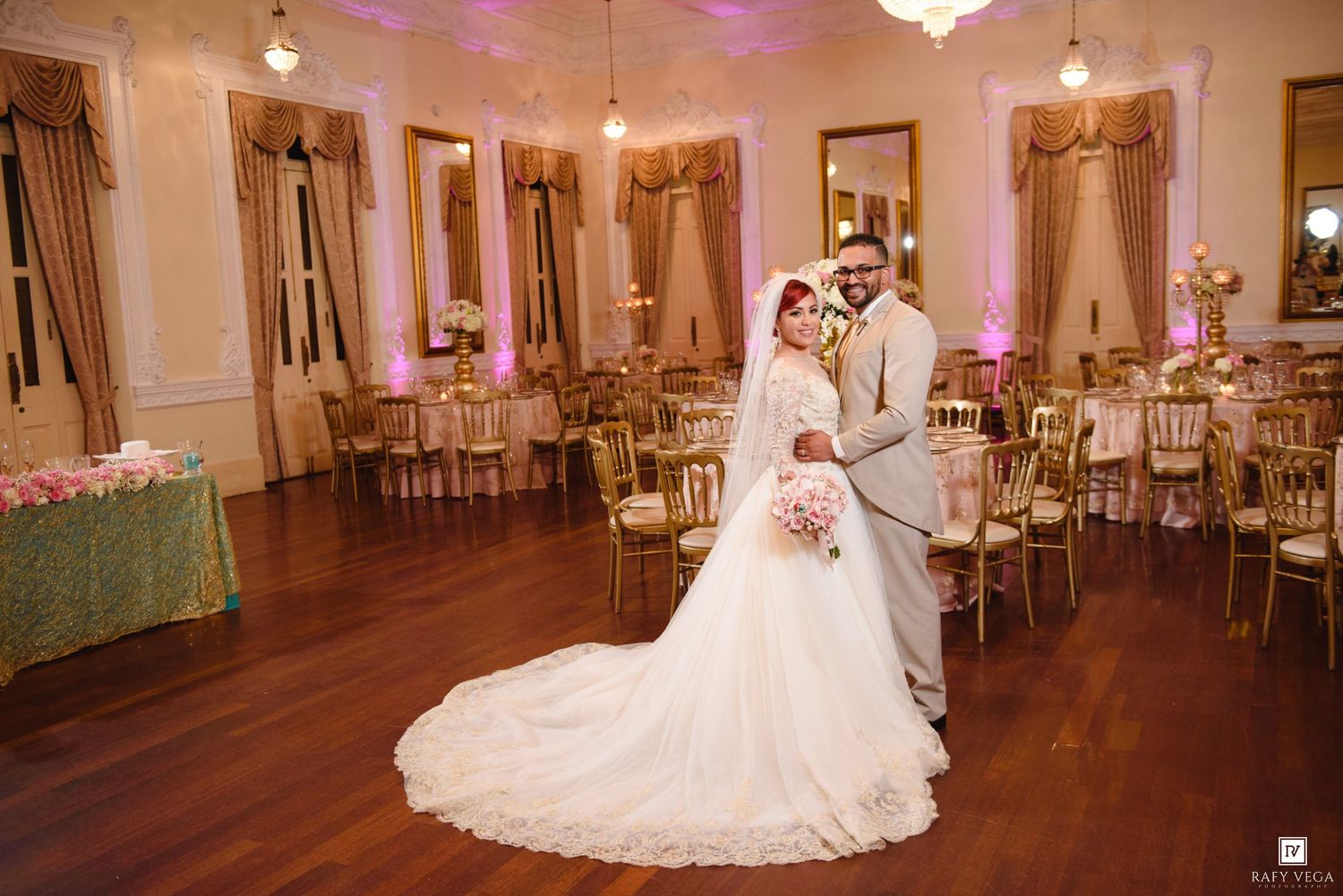 Bride and groom pose elegantly in a grand ballroom decorated with gold chairs and pink floral centerpieces for their wedding.