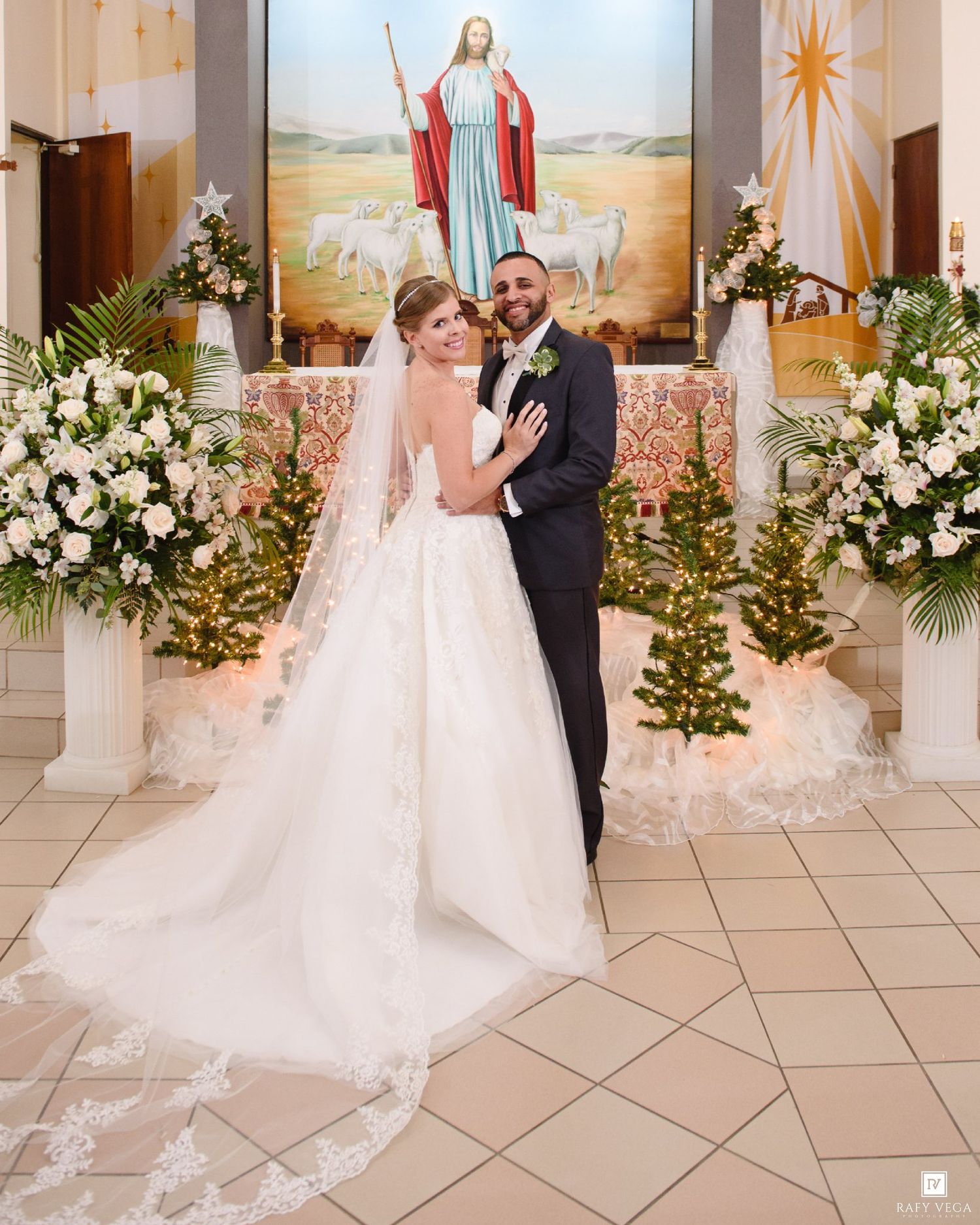 Bride and groom pose together at church altar adorned with white floral arrangements and Jesus painting backdrop.