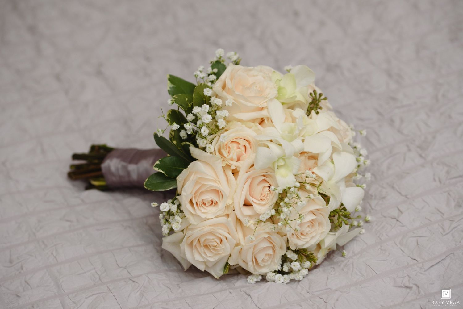 Elegant bridal bouquet of cream roses and baby's breath with green accents, tied with a gray ribbon.