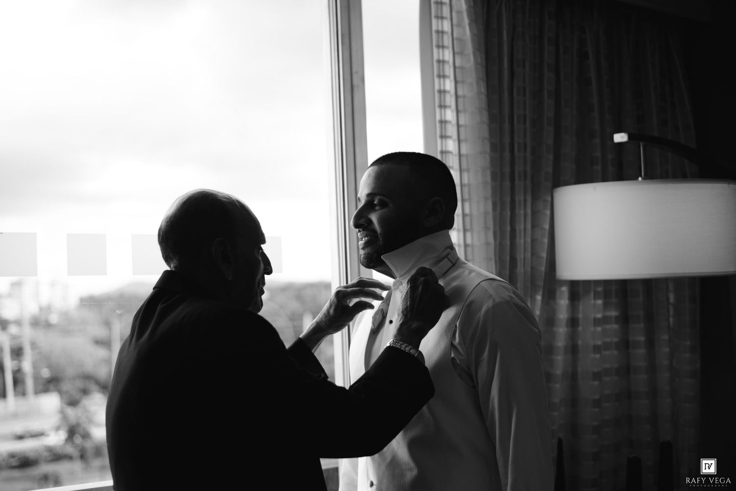 Black and white photo of a man helping another man adjust his tie near a window, likely a groom getting ready.