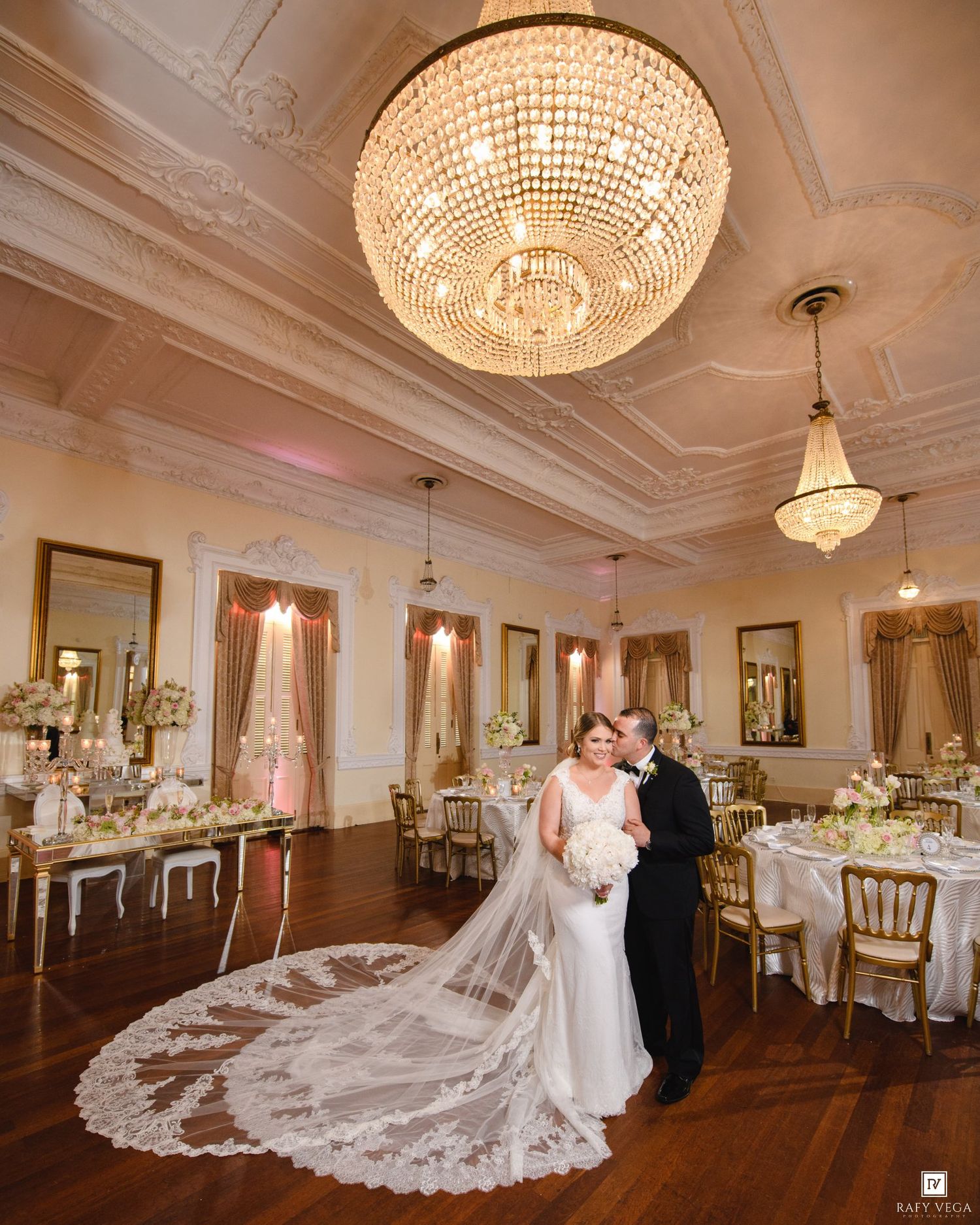 Bride and groom pose in elegant ballroom with crystal chandelier, floral arrangements, and lace cathedral veil.