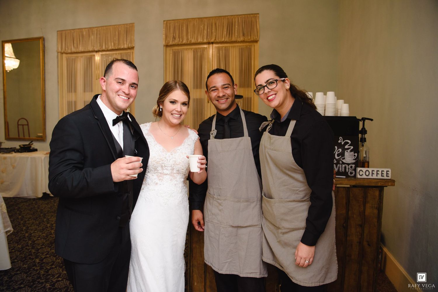 Newlywed couple poses with two catering staff members near a coffee station at their wedding reception.