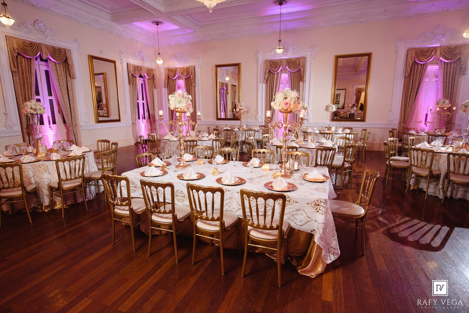 Elegant wedding reception hall with gold chairs, floral centerpieces, and romantic pink uplighting on white walls.