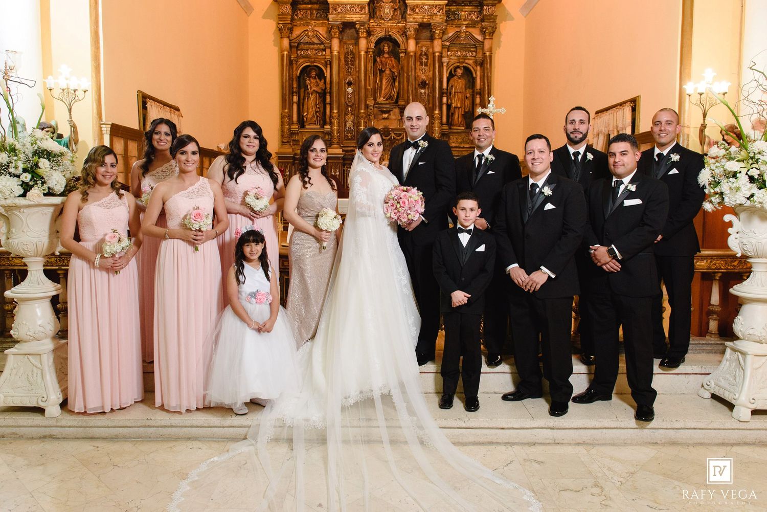 Wedding party poses in church with bridesmaids in blush pink gowns, groomsmen in black suits, and bride in white gown.