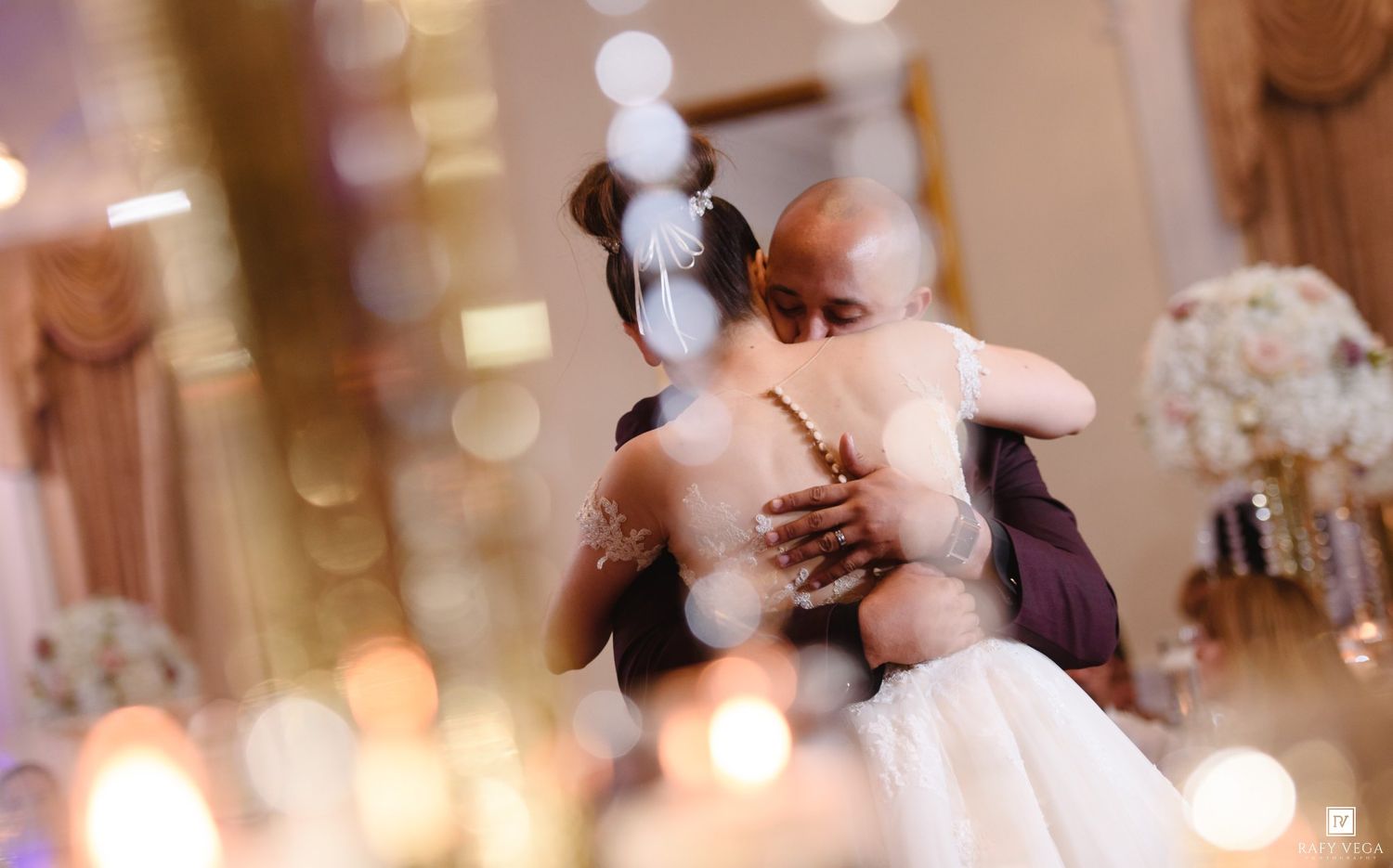 Couple sharing an emotional first dance embrace at elegant wedding reception with soft bokeh candlelight in foreground.